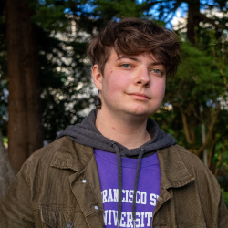 Elliot is smiling at the camera and wearing earrings that are large hanging bats. He is wearing a purple SF State T Shirt under a hooded jacket.
