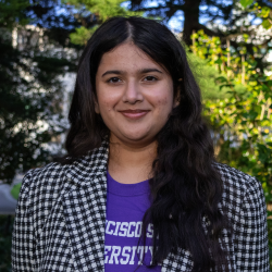 Divya, a girl with long wavy dark brown hair, is smiling. She is wearing a black and white gingham blazer over a purple SF State T Shirt.
