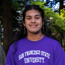 Sam has long brown hair and dangly earrings. He is wearing a purple SF State T Shirt.