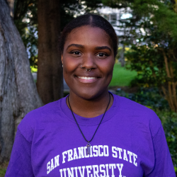 Eliza, a girl with a purple crystal necklace, is smiling. She is wearing a purple SF State T Shirt.