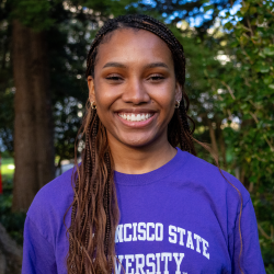 Christen Lewis, a person with long braided hair, is smiling. She is wearing a purple SF State T Shirt.