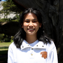 Emily is wearing a white SFSU shirt and standing in front of the SFSU Love tree
