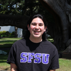 Giovanni is smiling, wearing a black SFSU shirt, standing in front of a tree