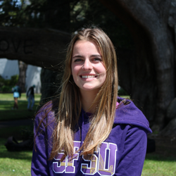 Lyric is smiling, wearing a purple SFSU sweater, standing in front of a tree
