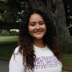 Naiseth is smiling, wearing a white SFSU shirt, standing in front of a tree