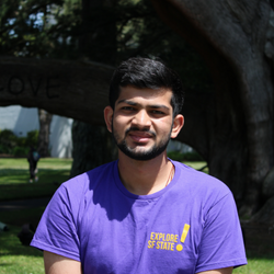 Shrey is smiling, wearing a purple SFSU shirt, standing in front of a tree