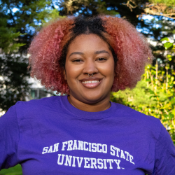 Kaylyn has curly pink hair and a septum piercing. She is smiling and wearing a purple SF State T Shirt.