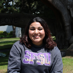 Xitali is smiling, wearing a grey SFSU sweater, standing in front of a tree